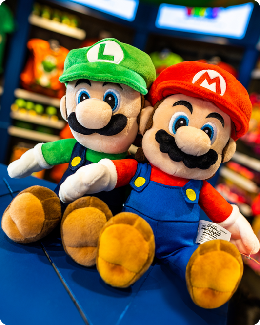 Plush toys of Mario and Luigi sitting on a blue surface with a store in the background - click to shop SUPER NINTENDO WORLD™ Drinkware now!